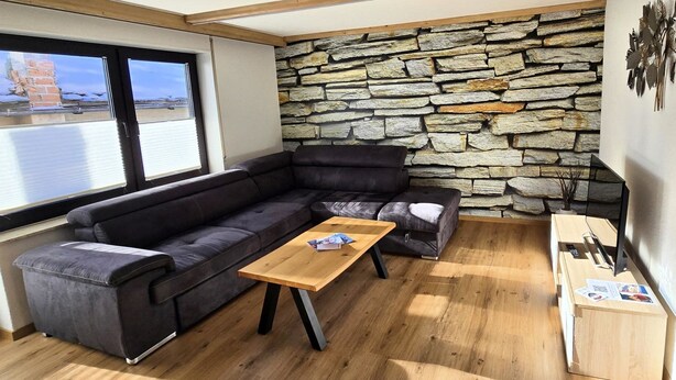 Un salon moderne avec un mur en pierre, un canapé en cuir noir, une table basse en bois et une vue sur l’extérieur.