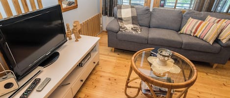Ein Wohnzimmer mit Flachbildfernseher, einer Couch, einem Korbsessel und einem Glastisch.