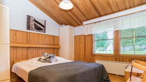 Un dormitorio con paneles de madera, una cama con manta gris, un almohadón, una silla y una ventana con persianas.