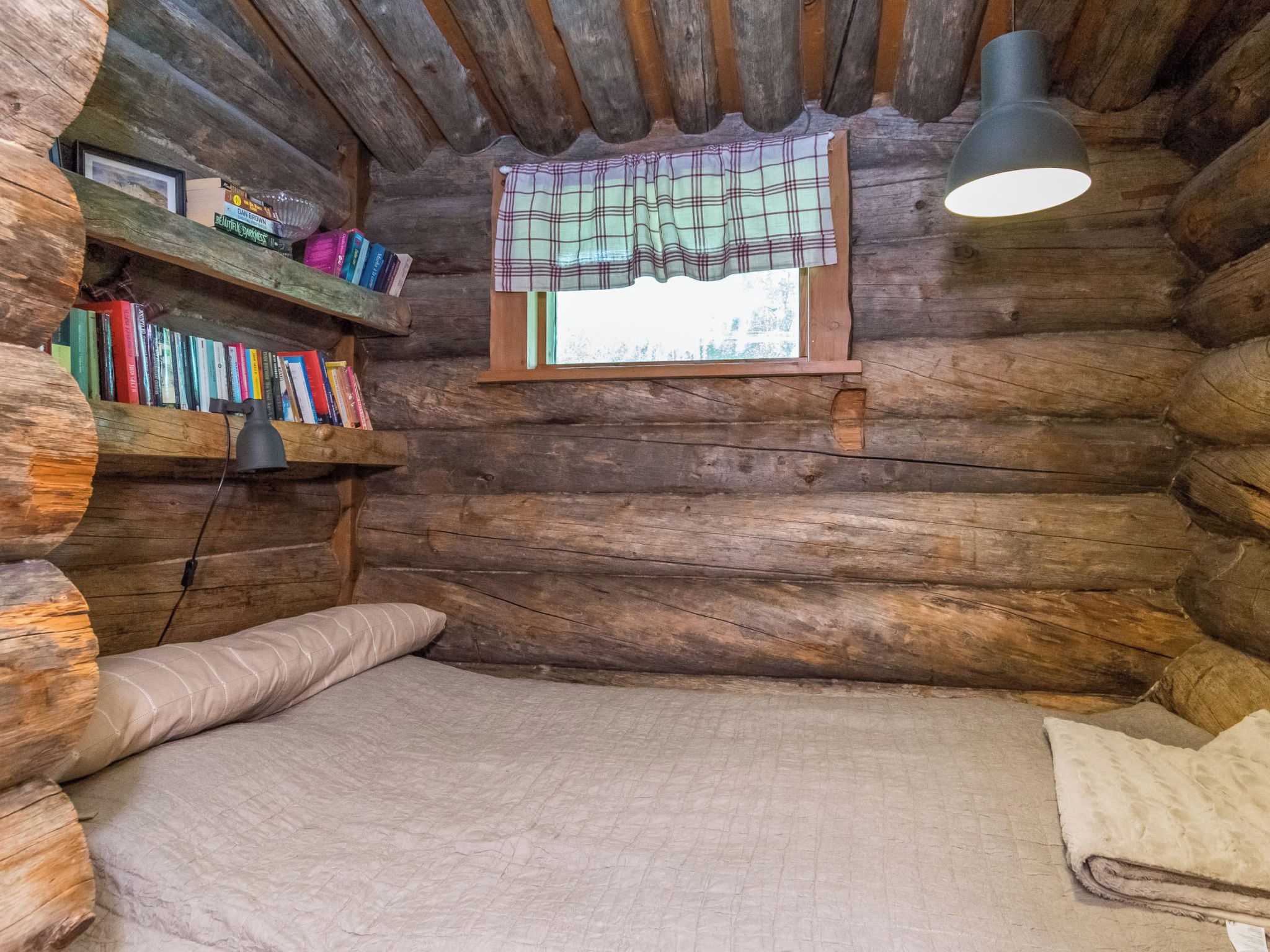 Une chambre dans un chalet en rondins, avec un lit, une fenêtre ornée de rideaux à carreaux, une étagère remplie de livres et un luminaire suspendu.