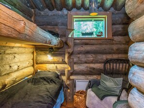 A log cabin room with a bed, a chair, a small table, and a window with a view of greenery.