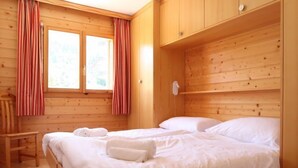 Ein Zimmer in einer Holzhütte mit zwei Betten, einem kleinen Holzstuhl und einem Fenster mit Vorhängen.