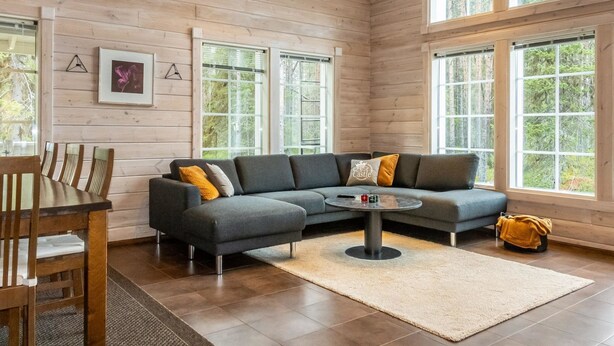 A modern living room with a sectional sofa, a dining table with chairs, and large windows offering a view of the forest.