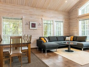 Et moderne interiør med træloft, spiseområde med stole og bord, en grå sofa med puder samt store vinduer.