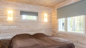 A wooden cabin bedroom with a bed, two wall-mounted lights, and a window with a view of trees.