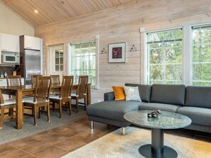 Una sala moderna con un sofá gris, sillas de madera, una mesa redonda y una cocina con refrigerador y microondas.