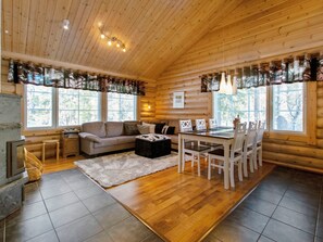 Interior de una cabaña de troncos con techo de madera, un sofá, una mesa de comedor con sillas y una chimenea.