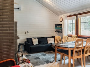A cozy living area with a black sofa, wooden dining table, and chairs, a flat-screen TV mounted on the wall, and a window with a view of trees.