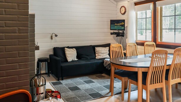 A cozy living room with a black sofa, wooden chairs, a TV, and a window with a view of trees.