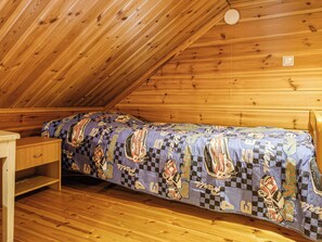 Un llit individual dins una cabana de fusta, amb un llençol de quadres i una petita taula de nit de fusta.