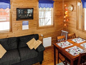 Ein gemütliches Cabin-Interieur mit Holzboden, einer schwarzen Couch mit Kissen, einem Esstisch mit Unterlagen und einer Uhr sowie einem Kalender an der Wand.