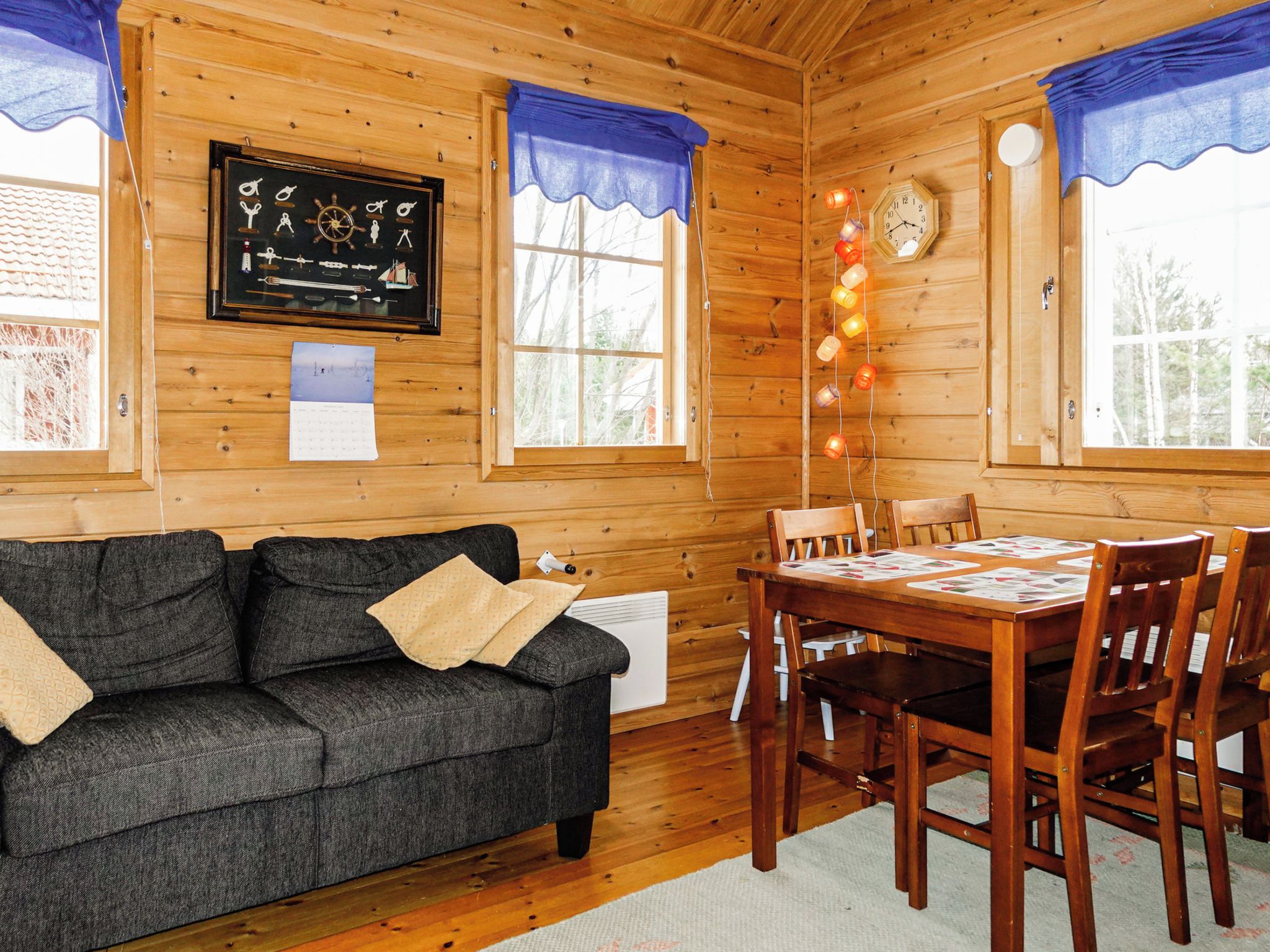 Interior acogedor de una cabaña con piso de madera, un sofá negro, una mesa de comedor con sillas y un reloj en la pared.