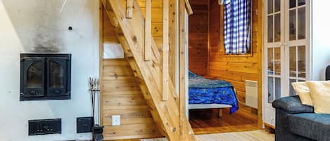 A cozy cabin interior with a wooden staircase, a bed with a blue blanket, a sofa, and a small heater.