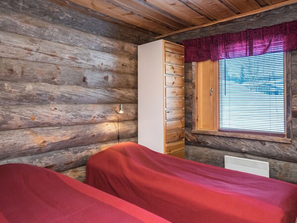 A cabin room with two beds, a wooden wall, a window with blinds, and a red curtain.