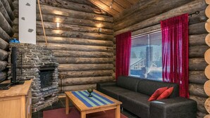 Ein Holzblockhaus-Interieur mit Kamin aus Stein, einer Couch, einem kleinen Tisch und einem Fenster mit roten Vorhängen.
