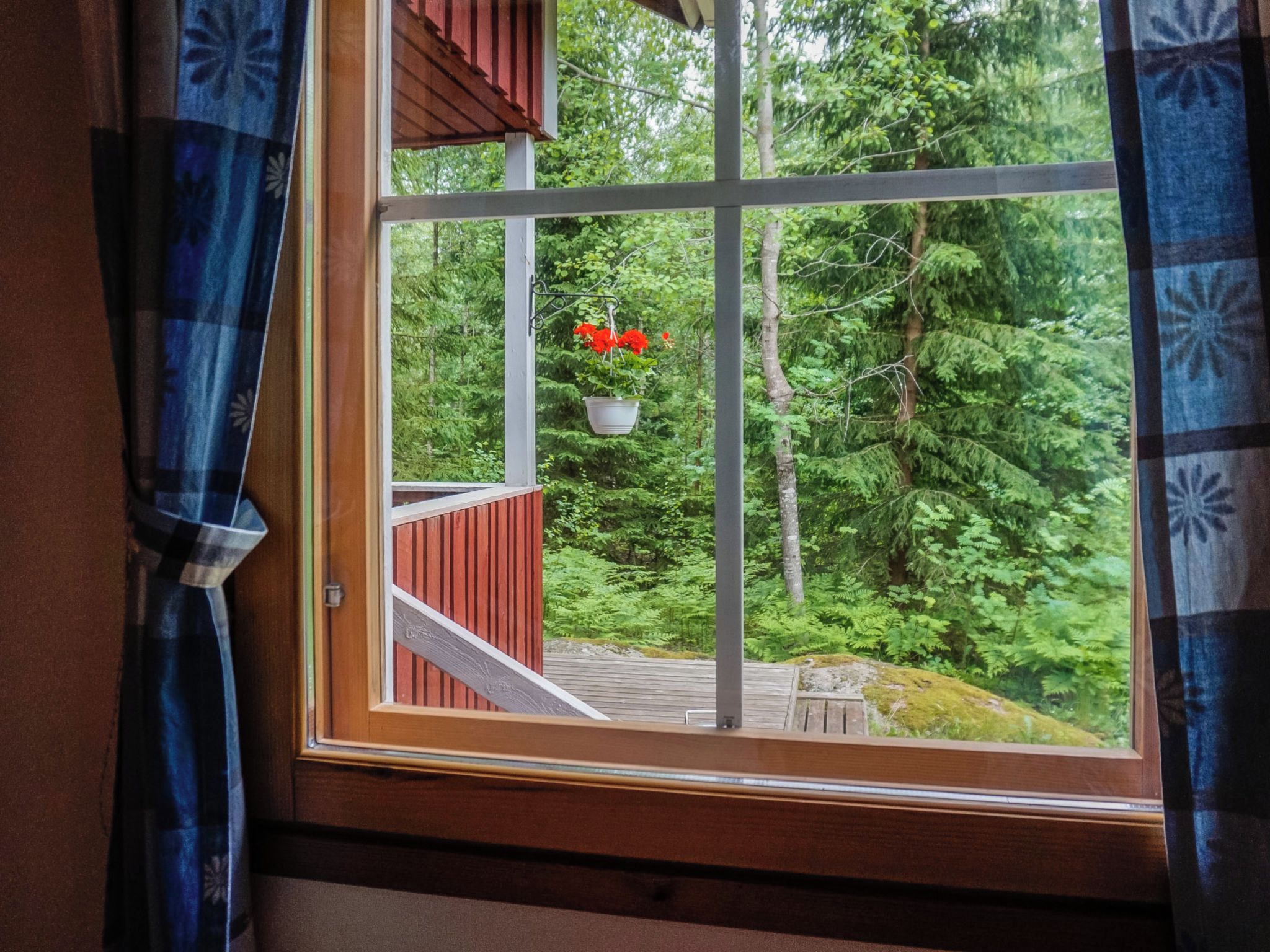 Une fenêtre donnant sur une terrasse en bois et ornée de pots de fleurs rouges suspendus.