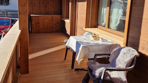 A wooden deck with a table and chair, a window with curtains, and a view of a building in the background. - Mösli (Zimmerli) by Interhome (Zweisimmen)