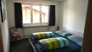 A small, single-bed room with a window, a radiator, a chair, and a radiator cover on the floor.
