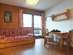 A cozy room with a wooden sofa, a dining table with chairs, a wooden floor, and a window with curtains.