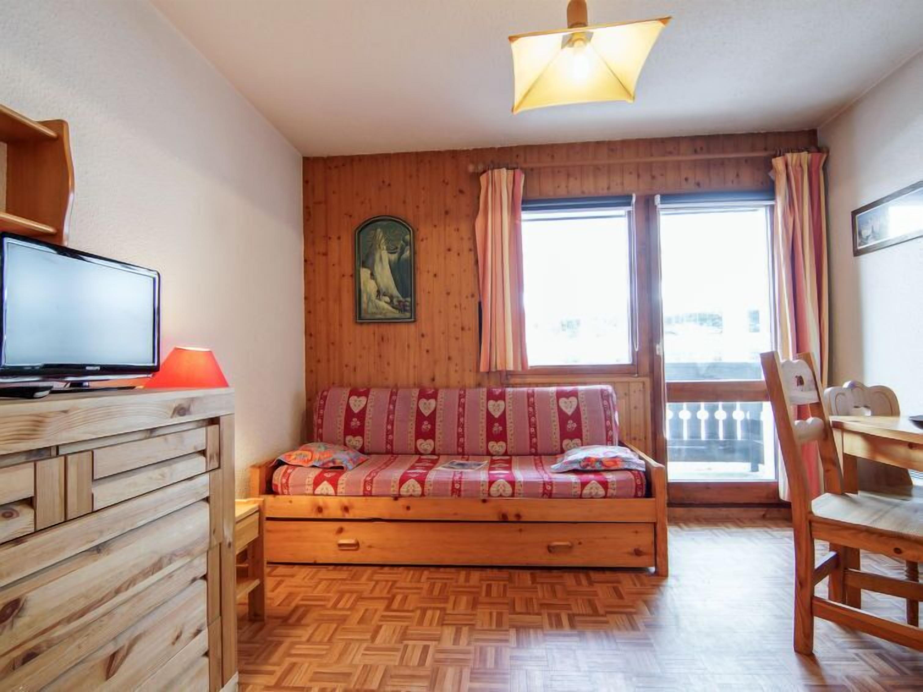 A cozy room with a wooden bed, a television on a wooden shelf, a dining table with chairs, and a window with curtains.