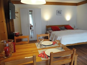 A room with a wooden dining table set for two, a bed with red and gray pillows, and a television mounted on the wall.