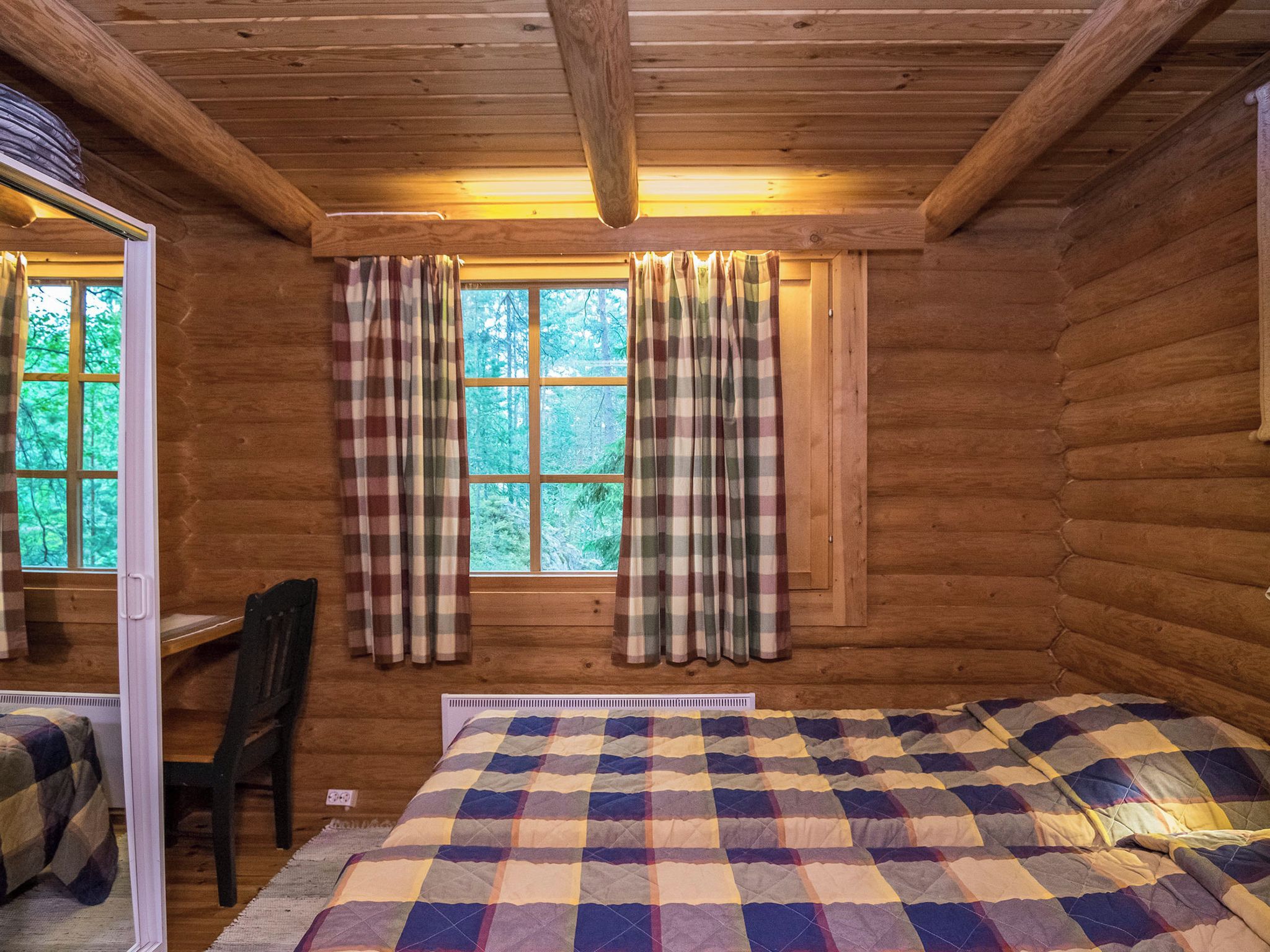 Ein Ferienhäuschen mit einem Bett, einem Sessel, einem Fenster mit Vorhängen und Blick auf den Wald.