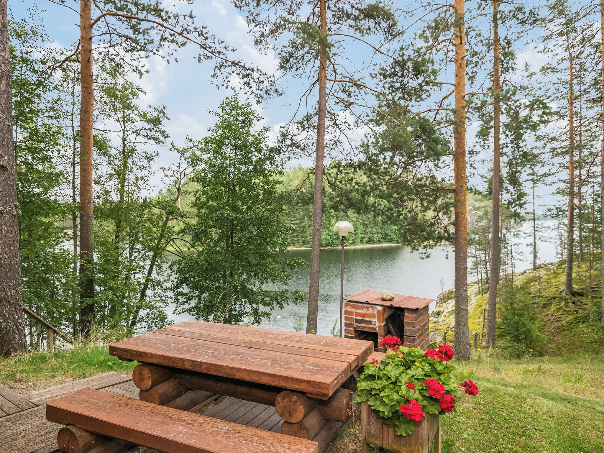 A wooden picnic table and a brick structure with a lamp post in a forested area near a lake.