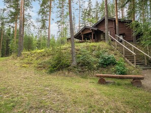 A rustic cabin with a wooden staircase leading up to a porch, surrounded by a forested area.