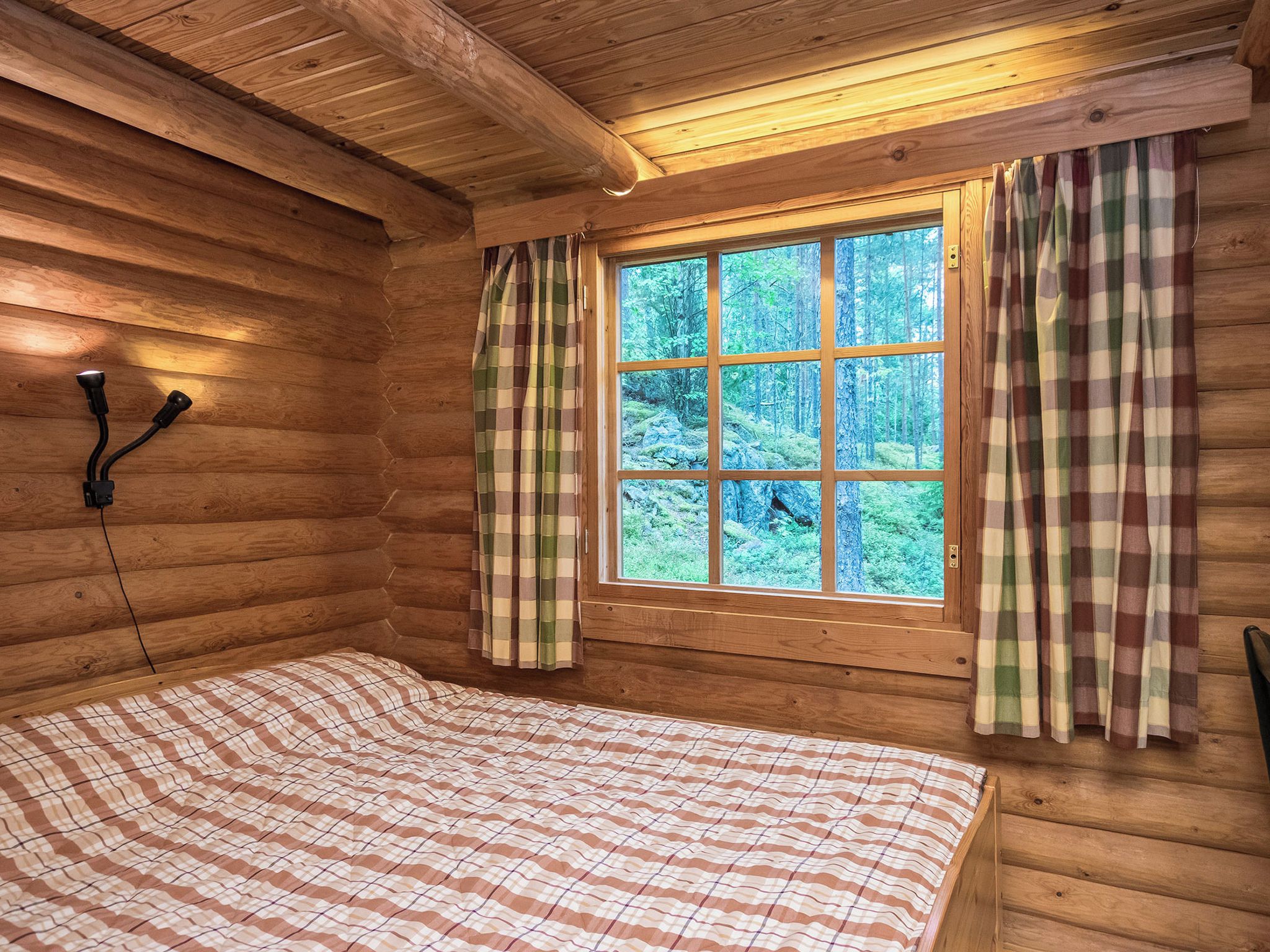 A wooden cabin room with a bed, a window with curtains, and a wall-mounted light fixture.