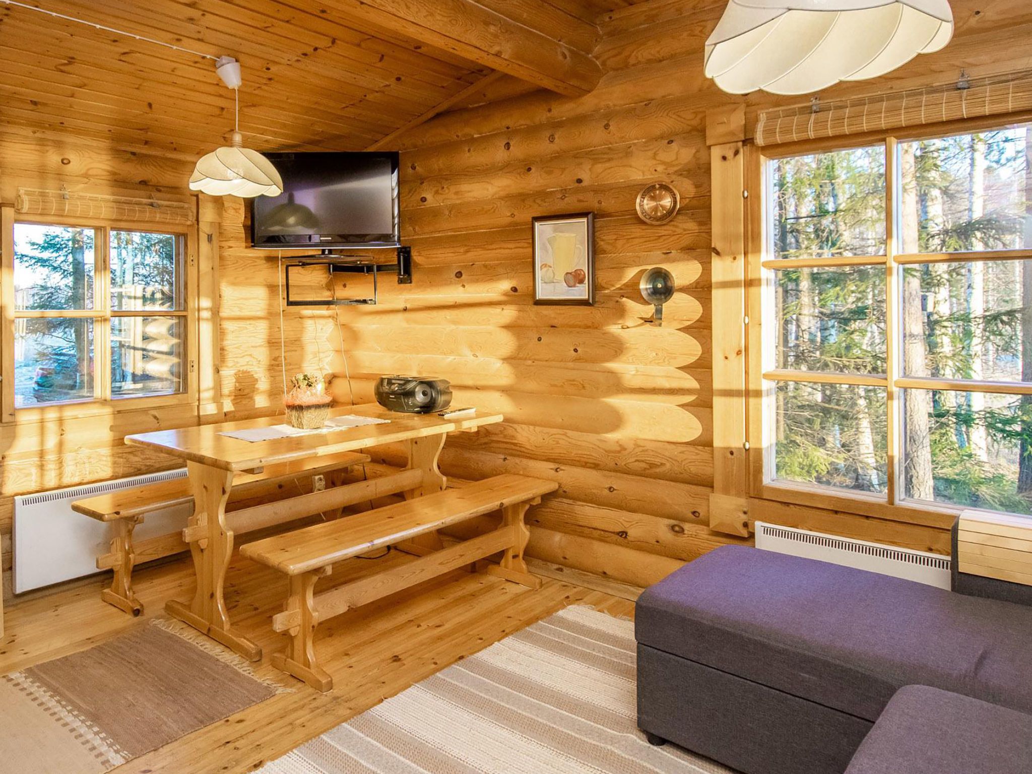 A log cabin interior with a wooden dining table, benches, a television mounted on the wall, and a large window offering a view of the forest.
