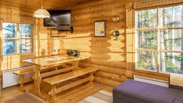 A log cabin interior with a wooden dining table, benches, a television mounted on the wall, and a large window offering a view of the forest.
