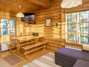 Ein Holzblockhaus-Interieur mit einem Holzesstisch, Bänken, einem an der Wand befestigten Fernseher und einem großen Fenster mit Blick auf den Wald.