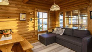 A cozy log cabin interior with a wooden dining table, a gray sofa with patterned cushions, and large windows offering a view of the outdoors.