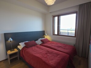 A hotel room with two beds, a nightstand, a lamp, a window with a view, and a radiator.