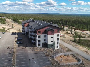 En bygning med flere etager, en parkeringsplads og et skovlandskab i baggrunden.