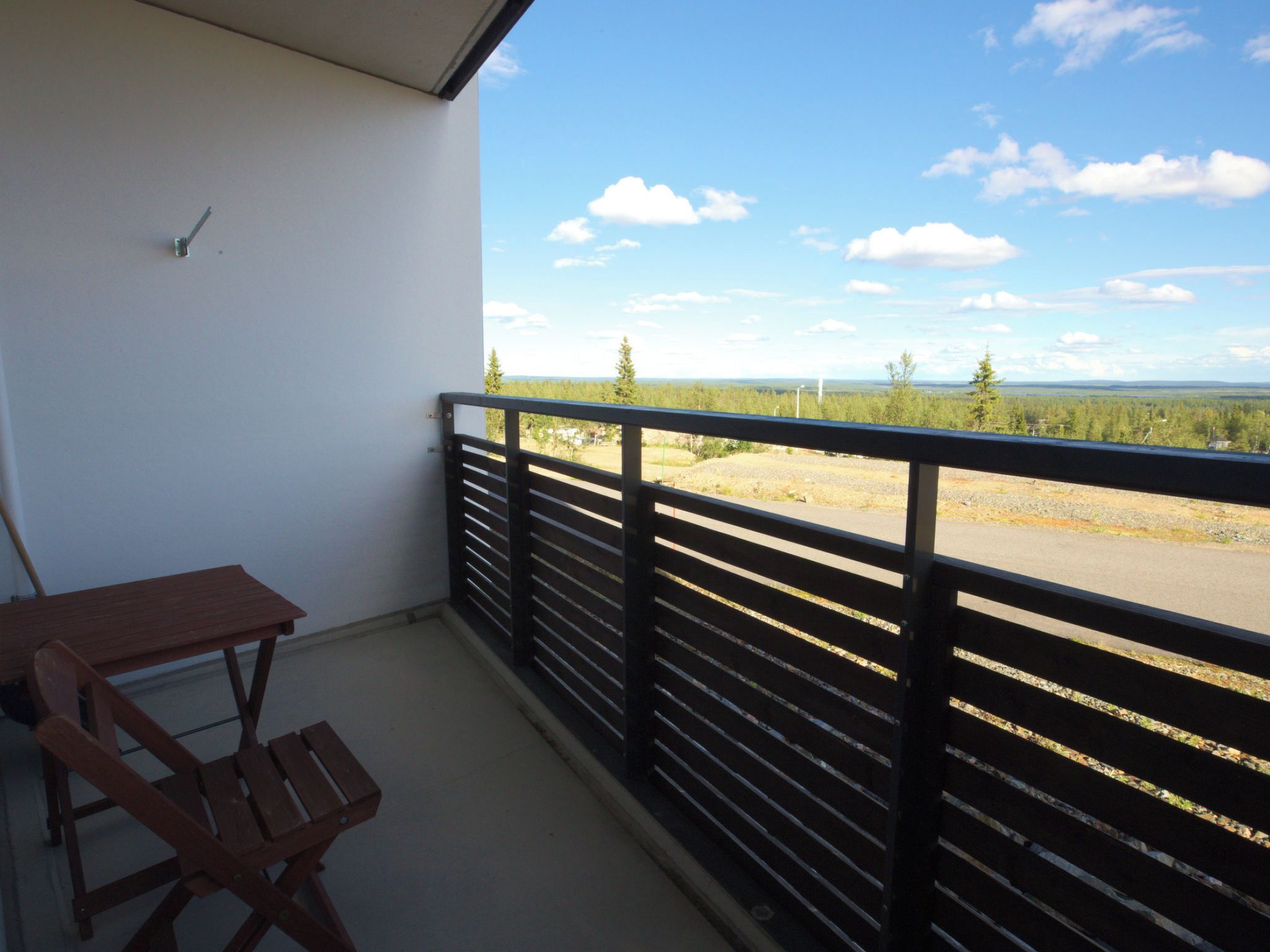 A balcony with a wooden table and chairs, a black railing, and a view of a forested landscape.