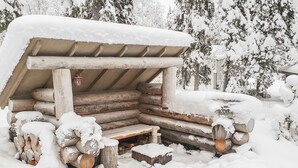 A snow-covered wooden cabin with a bench and logs piled outside.