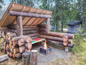 Een rustieke houten constructie met een schuin dak, rondom gestapelde boomstammen en een vuurkuil in het midden.