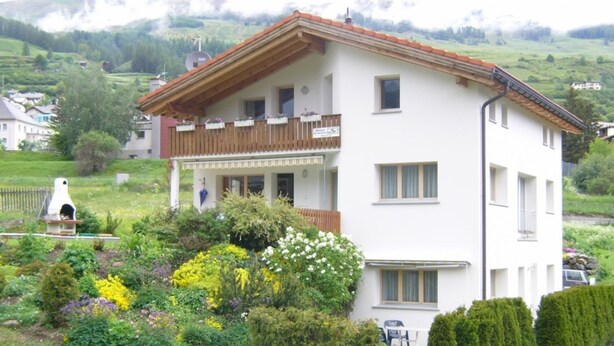 Un bâtiment à deux étages doté d’un balcon, entouré d’un jardin où poussent différentes plantes et fleurs.