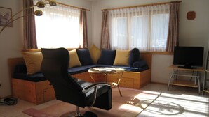 Un salon avec un canapé, un fauteuil, un téléviseur et une fenêtre avec des rideaux.