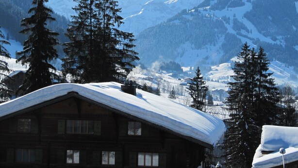 Eine Holzhütte mit schneebedecktem Dach, umgeben von hohen immergrünen Bäumen und schneebedeckten Bergen im Hintergrund.