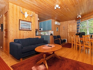 Interior de una cabaña de madera con sofá, mesa de comedor, sillas, zona de cocina y un ventanal grande.