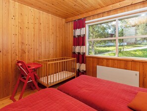 A room with wooden walls, a window with a view of greenery, a crib, and a bed with red bedding. - Valkeisrinne 1h by Interhome (Kuusamo)