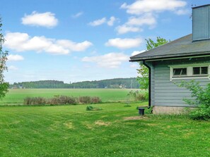 Ein Haus mit geneigtem Dach, einer kleinen Veranda und Blick auf ein grünes Feld und einen fernen Wald.
