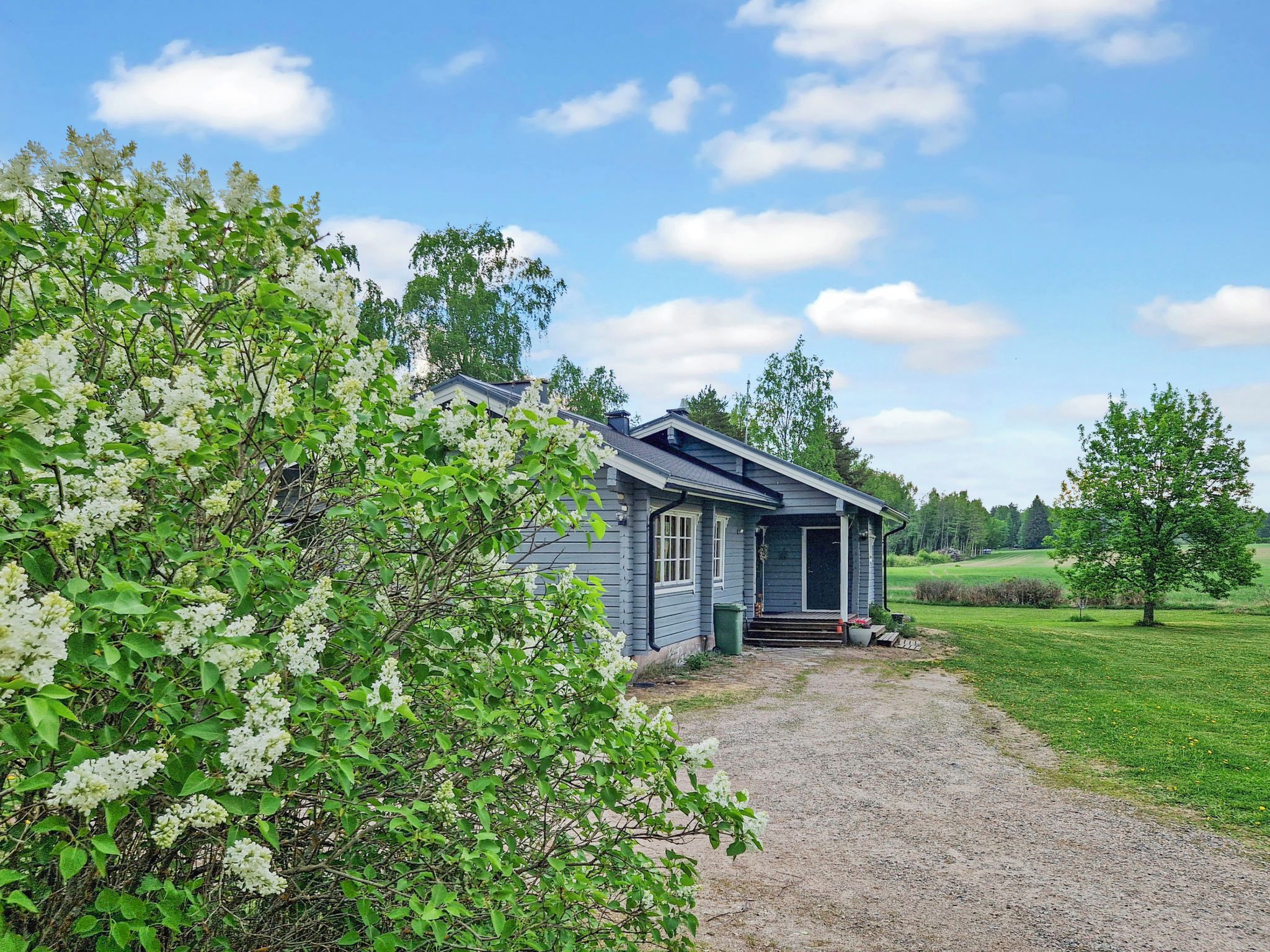 Ett litet hus i ett våningsplan med en veranda, omgivet av en grusuppfart och frodig grönska.