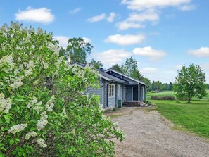 A small, single-story house with a porch, surrounded by a gravel driveway and lush greenery.