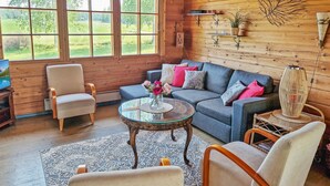 Interior acogedor de una cabaña con detalles de madera, un sofá gris con cojines coloridos, una mesa redonda de vidrio y vistas al follaje exterior.