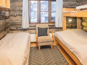 A log cabin room with two beds, a chair with a pillow, a window with curtains, and a view of snow-covered trees.