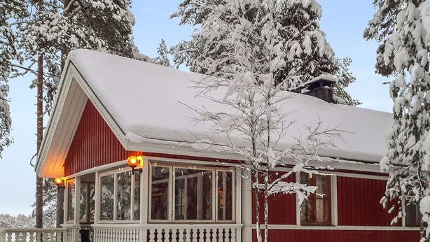 Une cabane en bois rouge, dotée d’un porche blanc et de grandes fenêtres, entourée d’arbres couverts de neige.