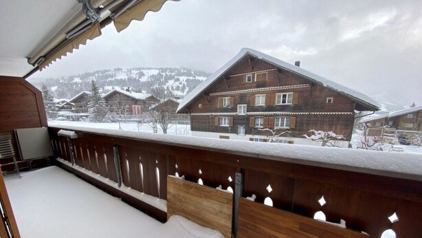 En træterrasse med sne på jorden og udsigt til en stor bygning med mange vinduer og et skråt tag.